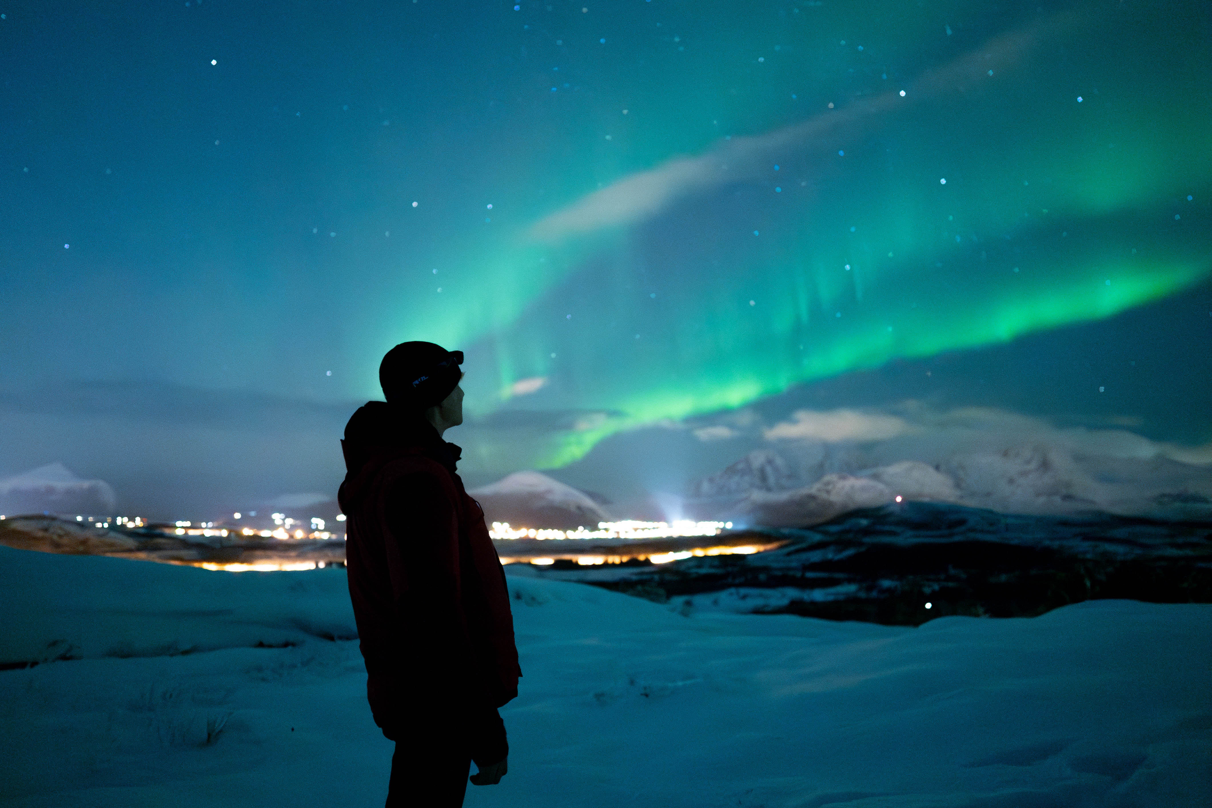 A figure looking up at the Aurora Borealis, with a distant city in the background